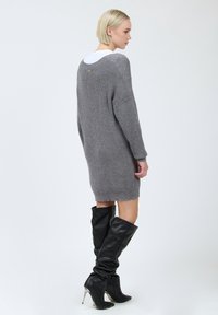 Graues Strickkleid mit lockerer Passform, weiten Ärmeln und rundem Ausschnitt. Details sind ein leicht ausgefranster Saum und ein metallischer Akzent auf der Rückseite.