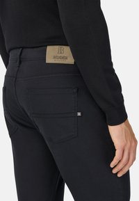 Jean de mezclilla negra con un diseño ajustado, que presenta un parche de cuero marrón claro etiquetado "BOGGI MILANO" y un pequeño detalle metálico en el bolsillo.