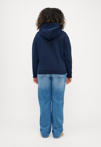Felpa blu navy con cappuccio regolabile, polsini a costine e una vestibilità ampia. Abbinata a jeans chiari a gamba larga con dettagli di cucitura in vista.