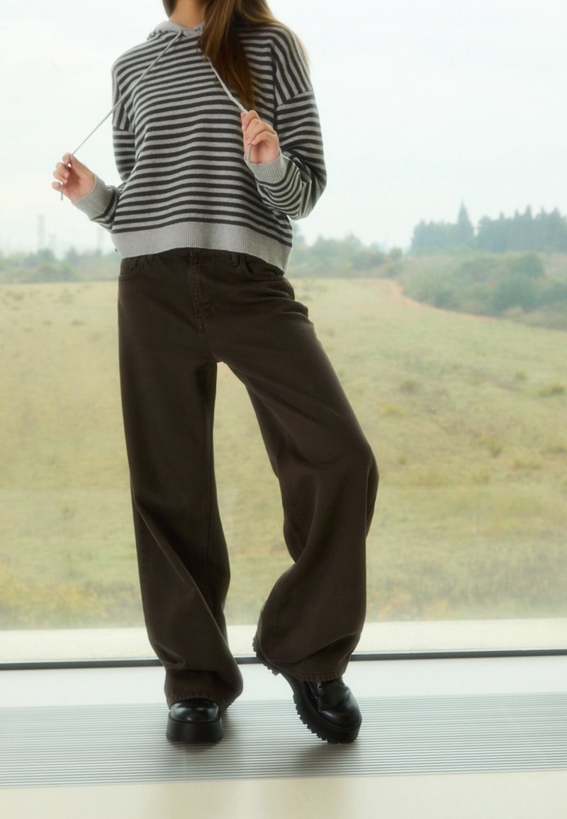 Personne portant des chaussures noires épaisses, un pantalon ample marron foncé et un sweat à capuche rayé gris et noir à manches longues, debout près d'une grande fenêtre.