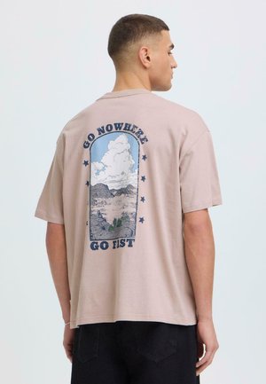 Jeune homme de profil portant un T-shirt beige avec un paysage de montagne et les textes "GO NOWHERE" et "GO FAST" au dos.