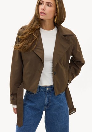 Femme portant une veste courte marron sur une chemise blanche avec un jean bleu, posant avec une main derrière le dos, regardant sur le côté.