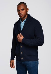Cardigan con colletto a scialle lavorato a maglia di colore blu navy, con tessuto testurizzato, cinque bottoni marroni e due tasche frontali, indossato sopra una camicia azzurro chiaro.