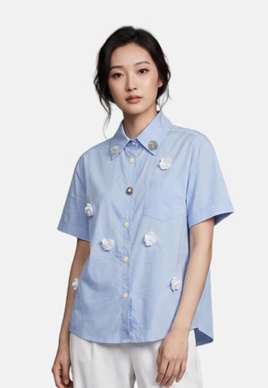 Femme portant une chemise bleu clair à manches courtes avec des appliqués floraux blancs et un pantalon blanc sur un fond uni.