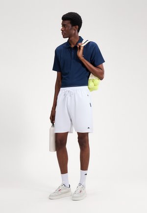 Homme en polo bleu marine et short blanc tenant une bouteille d'eau avec des balles de tennis dans un sac en filet sur l'épaule, debout et regardant vers la gauche.