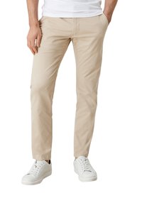 Homme portant un pantalon beige slim et des baskets blanches, debout avec une main dans la poche contre un fond blanc.