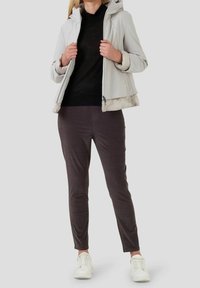 Žena oblečená v světle šedé bundě přes černý top, tmavě šedých kalhotách a bílých teniskách, stojící na jednobarevném pozadí.