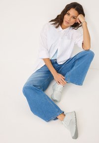 Jeans in denim azzurro chiaro a gamba larga, camicia bianca con colletto e sneakers grigie. Texture lisce e vestibilità rilassata. Modello seduto con un braccio appoggiato.