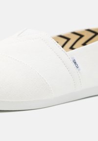 Witte canvas instapschoen met een gestructureerde bovenkant, voorzien van een zigzagpatroon op de binnenzool en een klein merkembleem aan de zijkant.