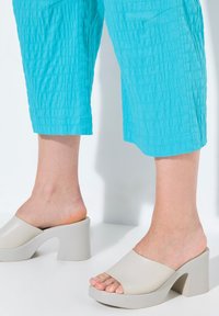 Mules beiges à talons épais avec un bout ouvert et une large sangle. Portés avec un pantalon turquoise court et texturé. Finition lisse, design moderne.