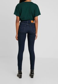 Mörkblå skinny jeans med hög midja, med ikoniska bakfickor och en tan läderlapp. Kombineras med en grön t-shirt med kort ärm.