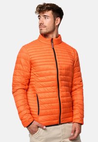 Orange quiltet jakke med høj krave, lynlåslukning fortil og sort lynlåselementer. Har horisontale syninger og sidelommer.