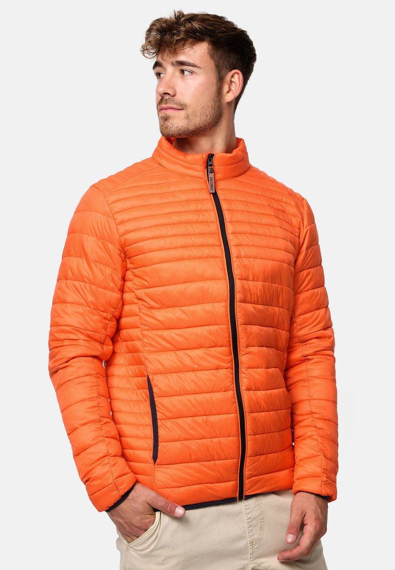 Orange quiltet jakke med høj krave, lynlåslukning fortil og sort lynlåselementer. Har horisontale syninger og sidelommer.