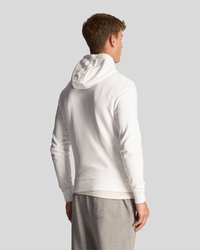 Haut en coton blanc avec capuche, manches longues et poignets côtelés. Porté par-dessus un t-shirt gris clair, associé à un pantalon de survêtement gris.