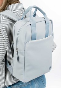 Ljusblå ryggsäck i slät material, med vadderade remmar, en frontficka och dragkedjestängning. Texturerade detaljer ger extra karaktär.