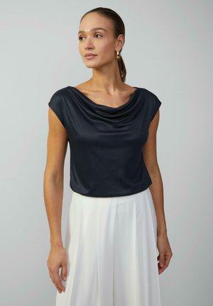 Top blu navy drappeggiato con maniche a cappuccio, realizzato in tessuto liscio. Abbinato a pantaloni bianchi a gamba larga, con pieghe per un tocco di texture in più.