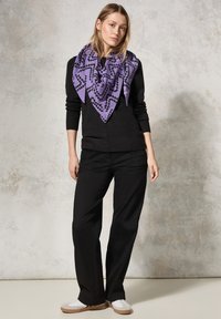 Sciarpa viola a motivi con design geometrico nero, indossata con un maglione nero, pantaloni neri e sneakers bianche, si staglia su uno sfondo neutro.