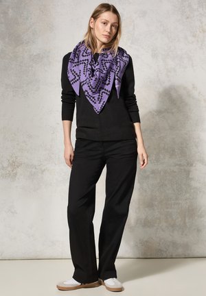 Sciarpa viola a motivi con design geometrico nero, indossata con un maglione nero, pantaloni neri e sneakers bianche, si staglia su uno sfondo neutro.