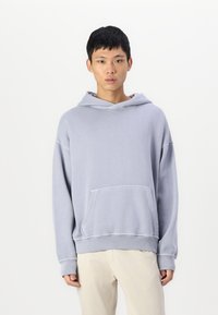 Grå oversized hættebluse lavet af blødt stof, med en frontlomme, snøre i hætten og ribbede manchetter, iført lyse bukser.
