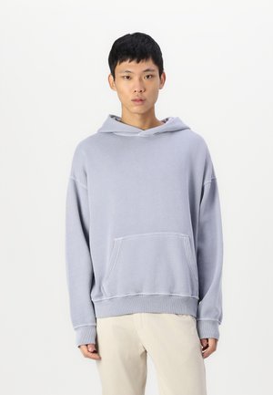 Hanorac supradimensionat gri, fabricat dintr-un material moale, având o buzunar frontal, glugă cu șnur și manșete ribbate, purtat cu pantaloni de culoare deschisă.