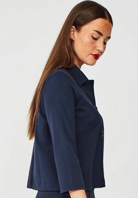 Navyblå tætsiddende jakke med krave, trekvartærmer og knapdetaljer. Glat overflade uden mønstre eller udsmykninger.