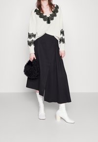 Weißer Pullover mit schwarzem Spitzenbesatz, schwarzer Wickelrock und weißen Stiefeletten mit Absatz. Hält eine schwarze flauschige Handtasche.
