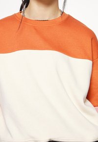 Osoba nosící oranžovo-bílé tričko s blokováním barev, u krku viditelný řetízkový náhrdelník.