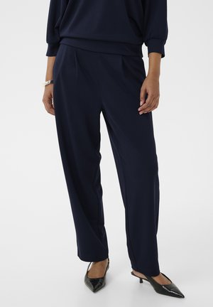 Woman wearing navy blue straight-leg pants, black pointed-toe slingback heels, and a matching navy top, standing against a plain background.