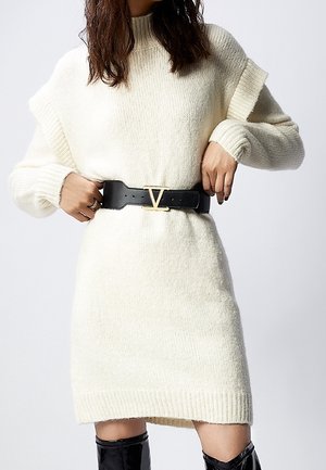 Vrouw die zwarte brede riem met gouden gesp over crèmekleurige gebreide sweaterjurk verstelt, gekleed in zwarte laarzen tot aan de knie tegen een effen achtergrond.