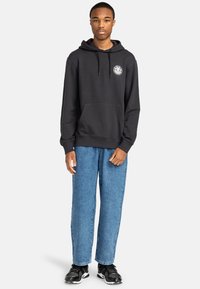 Black hoodie with a kangaroo pocket and logo on the chest, paired with light blue straight-leg jeans and black athletic shoes.