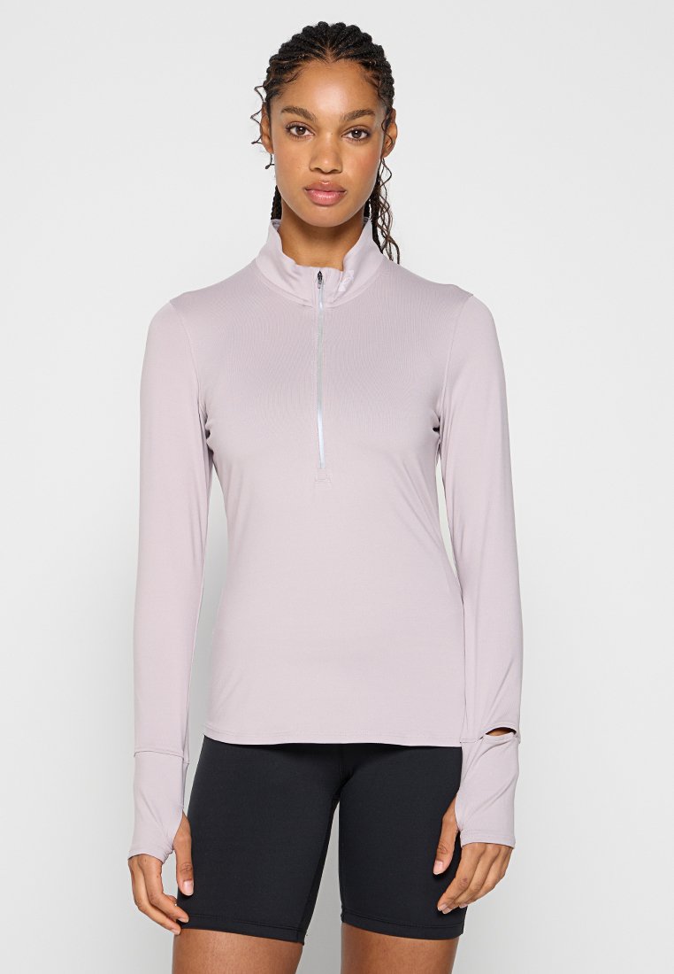 Femme avec des cheveux tressés portant un haut zippé à manches longues rose clair et un short de cyclisme noir, se tenant devant un fond uni clair.
