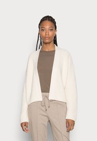 Cardigan creme de malha canelada com frente aberta, mangas longas e um corte relaxado, combinado com um top castanho justo e calças bege com cordão.