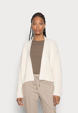 Cardigan creme de malha canelada com frente aberta, mangas longas e um corte relaxado, combinado com um top castanho justo e calças bege com cordão.
