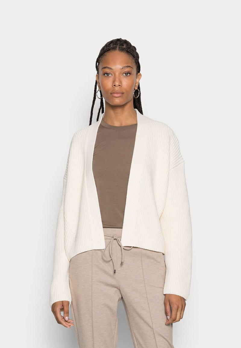 Cardigan creme de malha canelada com frente aberta, mangas longas e um corte relaxado, combinado com um top castanho justo e calças bege com cordão.