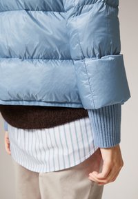 Light blue puffer jacket featuring a shiny, smooth texture; thick, quilted sections; and oversized sleeves, layered over a brown sweater and striped blouse.