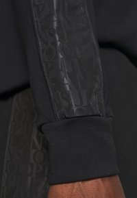 Černá mikina vyrobená z hladkého materiálu, s lesklým černým bočním panelem s vyraženým textem loga. Rukávy jsou žebrované a elastické.