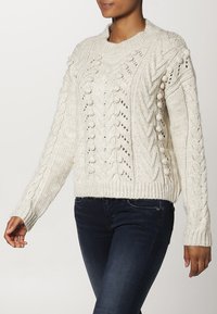 Gruby kremowy sweter z dzianiny z warkoczowymi i kuleczkowymi wzorami, okrągły dekolt, ściągacze na mankietach i u dołu, zestawiony z ciemnymi jeansami.