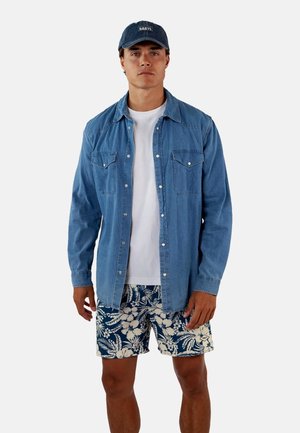 Denimshirt in een lichtblauwe kleur, met twee borstzakken, gelaagd over een wit T-shirt. Gecombineerd met marineblauwe shorts met bloemenprint.
