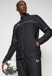 Veste de jogging noire zippée avec des rayures blanches, assortie à un pantalon ; dotée d'un col montant et de poches latérales. Tiens un ballon de football noir et blanc.