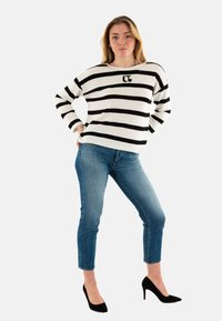 Maglione a righe bianco e nero con scollo rotondo e logo, abbinato a jeans slim blu e tacchi alti neri.
