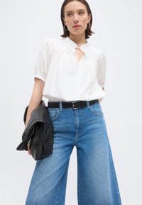 Γυναίκα που φοράει λευκό πουκάμισο και φαρδιά μπλε τζιν, κρατώντας ένα μαύρο υφασμάτινο σακάκι, στέκεται μπροστά σε ένα απλό ανοιχτό φόντο.