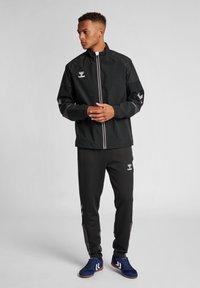 Schwarze sportliche Jacke mit grauen Akzenten, Reißverschluss vorne und Logo. Getragen mit schwarzen Hosen mit seitlichen Streifen und blauen Sportschuhen.