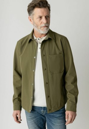 Muž so sivou bradou, oblečený v olivovo zelenej košeľovej bunde na gombíky cez bielu henley košeľu a modré džínsy, hľadí do strany.