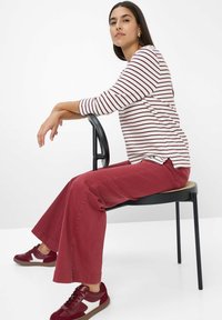 Gestreiftes Langarmshirt mit roten und weißen Streifen, kombiniert mit hochgeschnittenen roten Hosen und bordeauxfarbenen Turnschuhen. Modell sitzt auf einem schwarzen Stuhl.