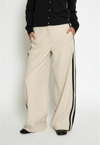 Beige wide-leg trousers with black side stripes, featuring side pockets and a smooth, lightweight fabric. Paired with a black button-up cardigan.
