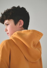 Persona joven con cabello rizado oscuro vistiendo una sudadera naranja, mirando hacia la izquierda contra un fondo claro y liso.
