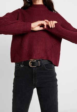 Woman wearing a maroon knit sweater and black jeans with a black belt, adjusting her sleeves against a plain light background.
