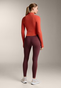 Rashguard in rood met lange mouwen, naadloos ontwerp. Gecombineerd met burgundy leggings met een zijstreep en een aangesloten silhouet.