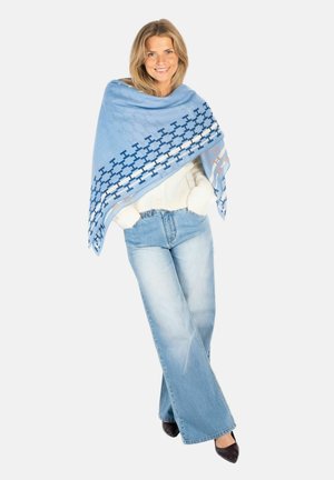 Lachende vrouw met blond haar draagt een lichtblauwe sjaal met patroon, een witte trui, wijde jeans en zwarte puntige schoenen, poseert tegen een witte achtergrond.