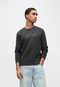 Hosszú ujjú fekete texturált pulóver kerek nyakkivágással és finom, a mellkasnál elhelyezett nyomott logóval, világoskék farmernadrággal kombinálva.